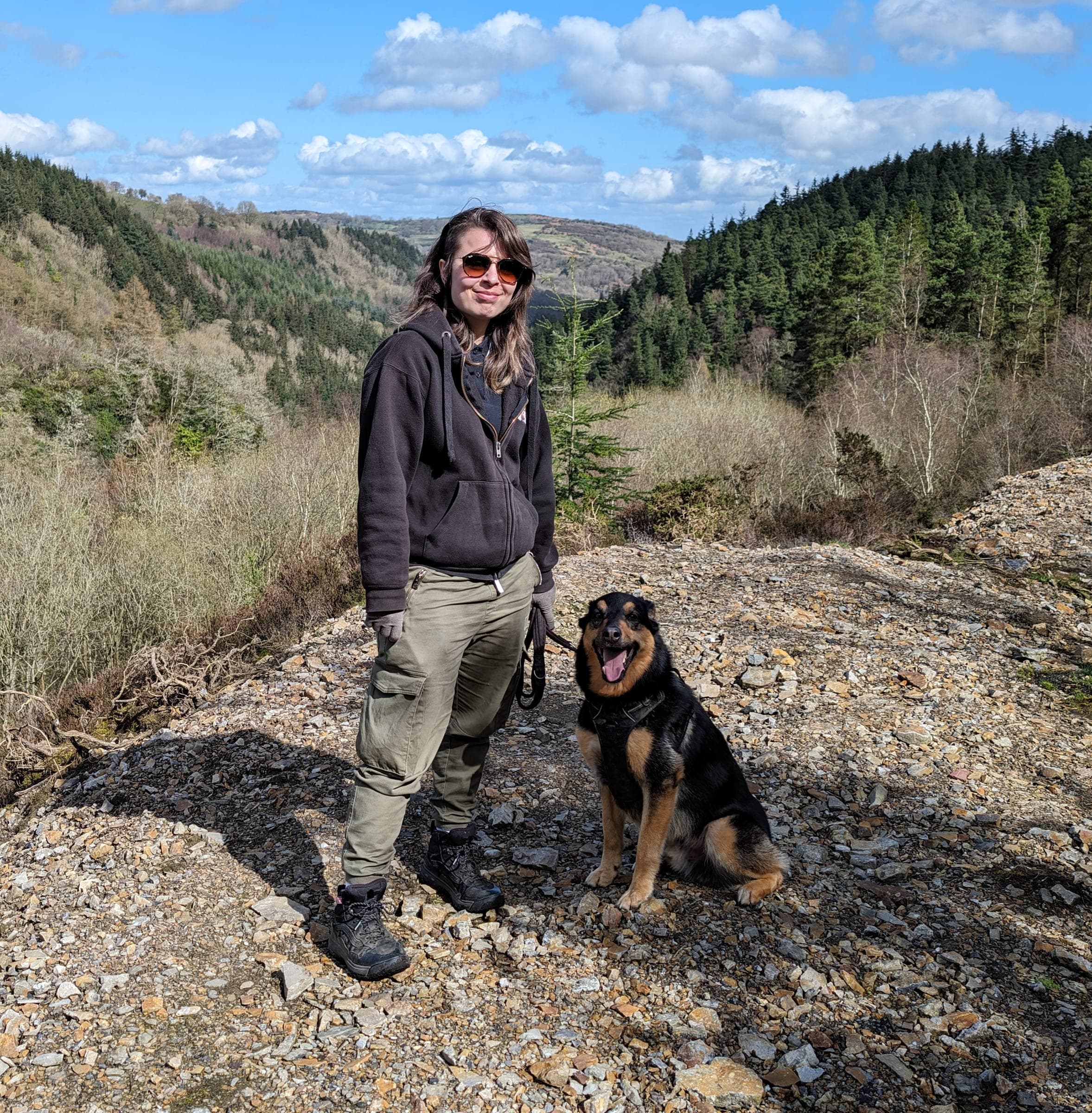 Senay from Fairy Dog Mothers standing on a hilltop trail with a happy black and tan dog sitting beside her